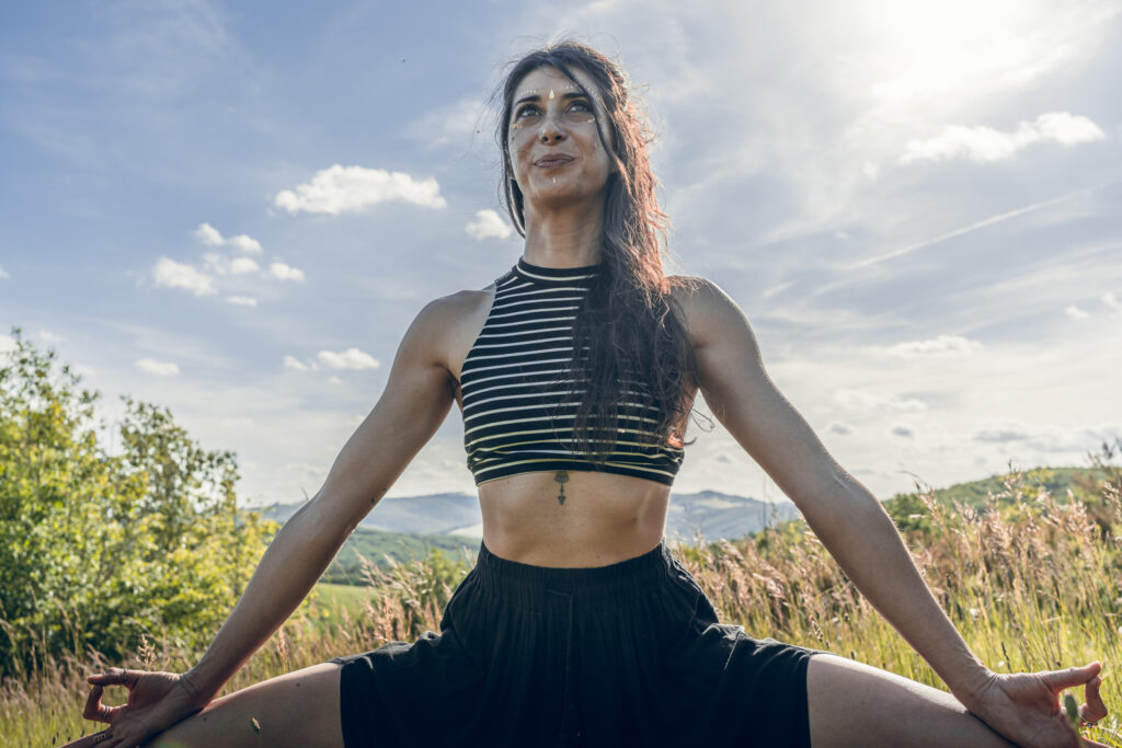 À propos Marylou Forges, fondatrice de Yoga Le Refuge à Saint-Rome-de-Tarn, en posture de la déesse dans un champ ensoleillé avec vue sur les collines de l’Aveyron