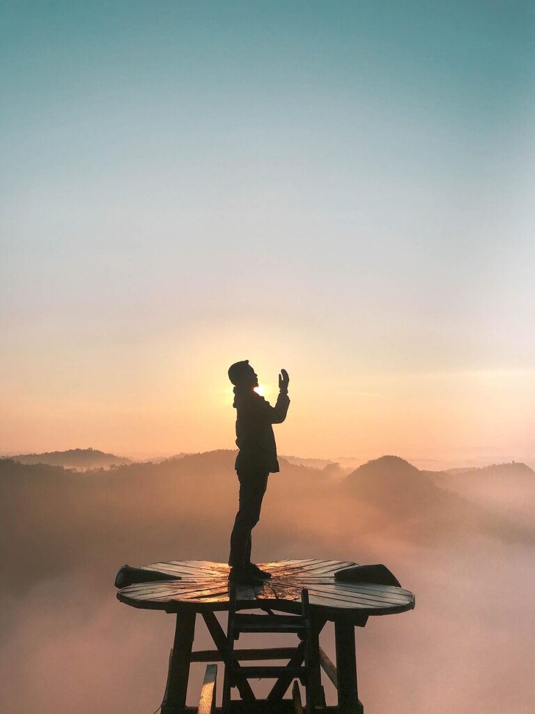 Silhouette d’une personne mains vers le ciel au lever du soleil, debout sur une plateforme en bois face aux montagnes et à la brume.