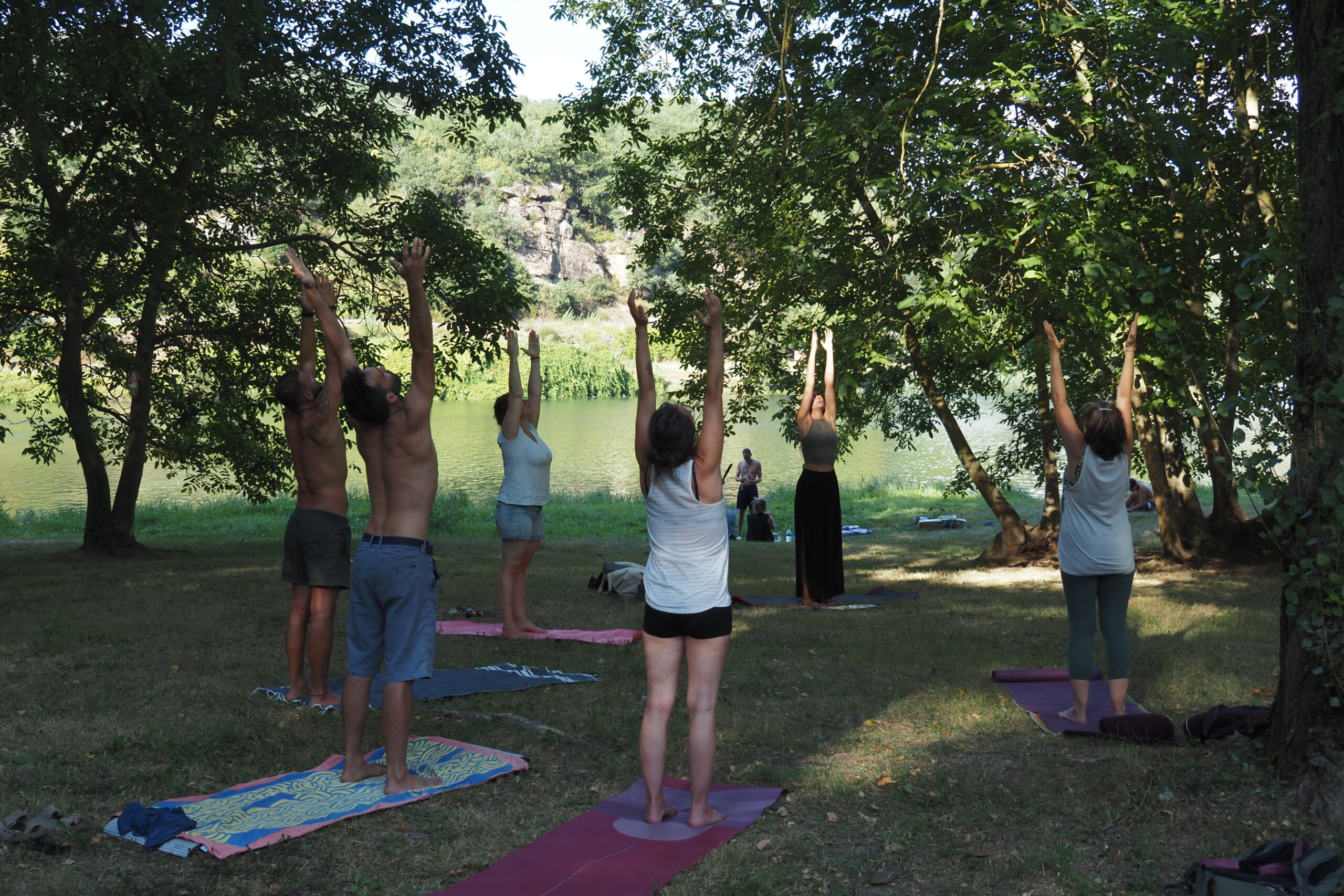 Petit groupe mixte en cercle lors d’un cours de yoga en plein air, bras levés vers le ciel dans la salutation au soleil.