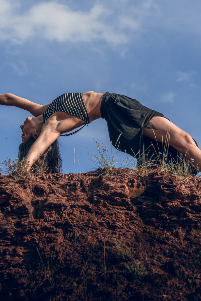 Posture de yoga courbée en arrière devant un ciel bleu avec des nuages, en équilibre sur une formation rocheuse texturée.