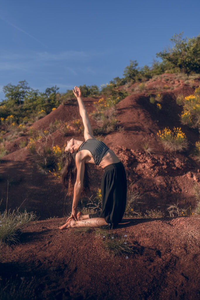 Posture du Chameau, bras tendu vers le ciel, sur une terre rouge en extérieur.
