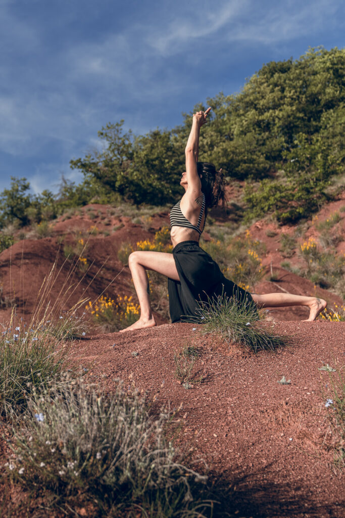 Marylou en posture du Croissant de Lune (Anjaneyasana) dans un paysage naturel rouge et végétalisé sous un ciel bleu.