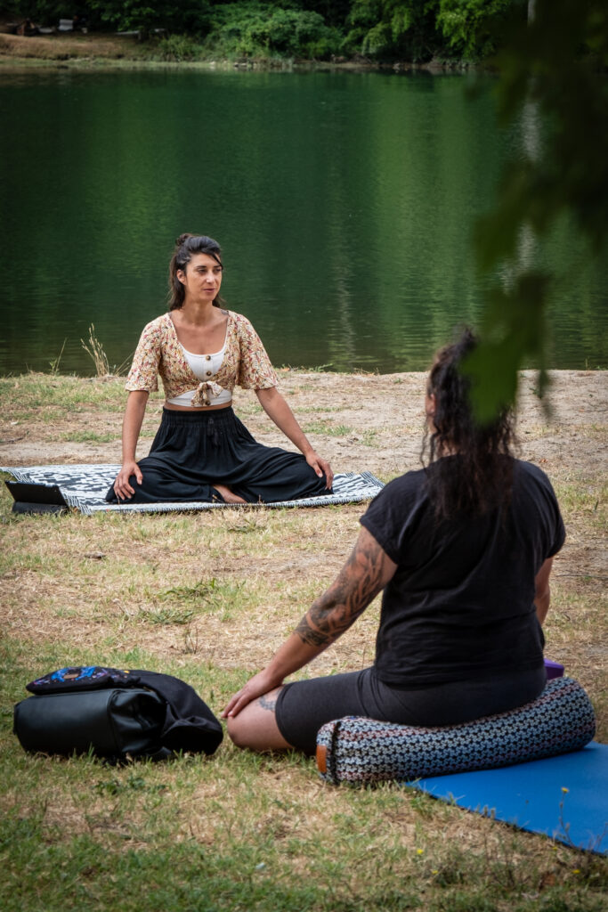 Séance de yoga en plein air au bord d’un lac, deux femmes assises en méditation sur leur tapis face à l’eau.