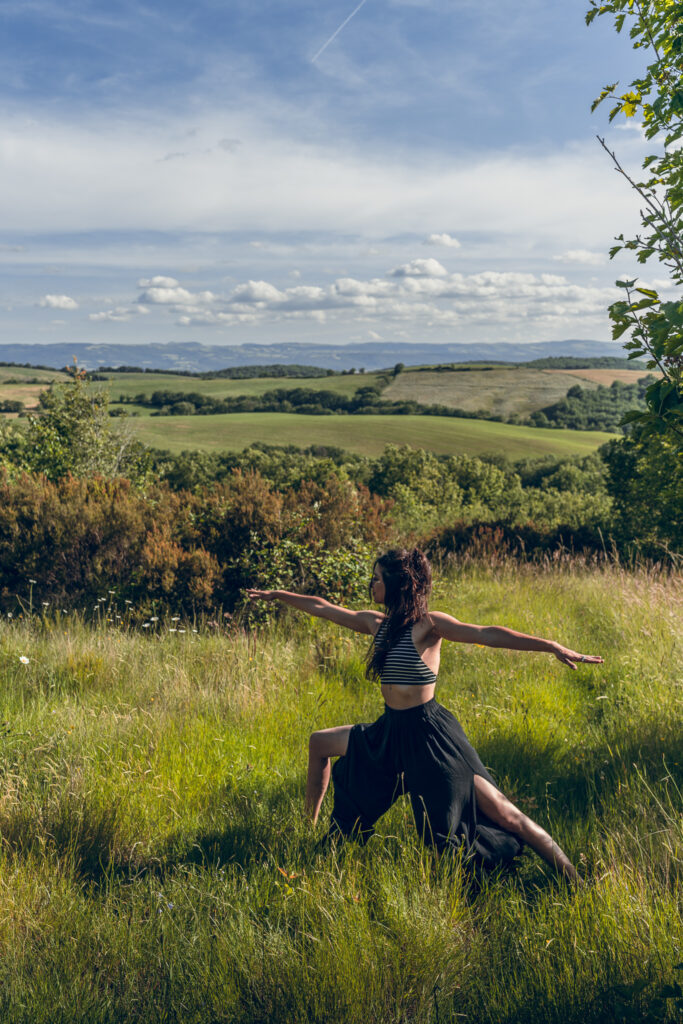 Photographie de la posture du Guerrier 2 (Virabhadrasana II) dans une prairie verdoyante sous un ciel bleu, symbolisant la force et l'élément feu.