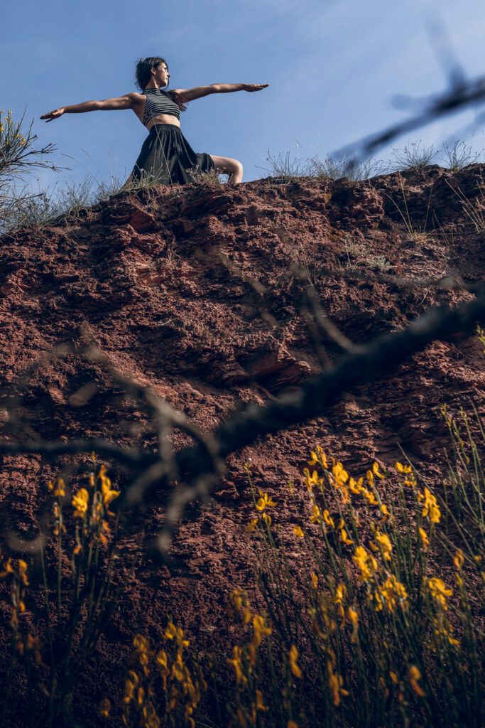Femme pratiquant la posture du Guerrier II (Virabhadrasana II) au sommet d'une falaise de terre rouge volcanique. Yoga en plein air axé sur l'ancrage et la force intérieure.