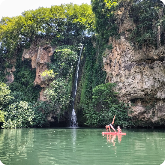 Marylou Forges en posture de yoga en équilibre sur un paddle board (SUP) avec son chien, sur une eau calme devant la cascade des Baumes, entourée par des falaises rocheuses et une végétation verdoyante.
